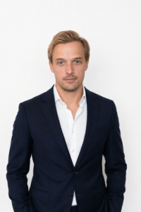Porträtt av Carl C G Eriksson. Han har kort blont hår, är klädd i en marinblå kostym och en vit skjorta, står framför en enfärgad vit bakgrund och tittar in i kameran.
